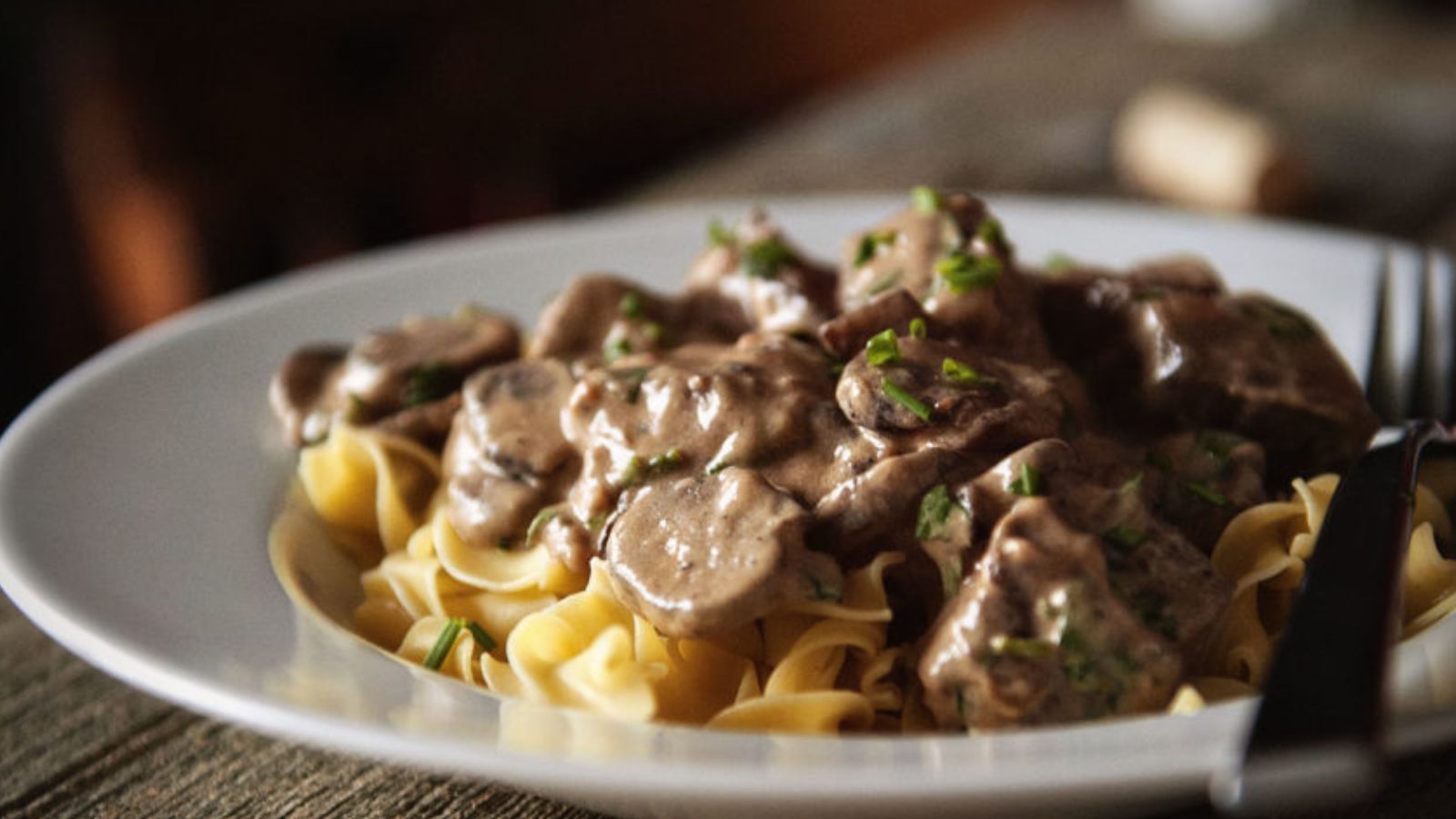 A plate of pasta topped with a creamy beef and mushroom sauce, garnished with chopped herbs, next to a fork.