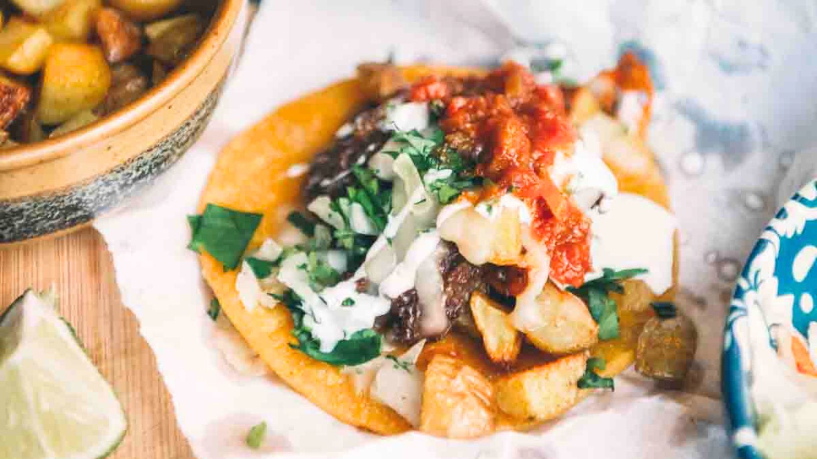 Taco with diced potatoes, black beans, cheese, cream, red salsa, and chopped cilantro. Lemon wedge and bowl of potatoes nearby.