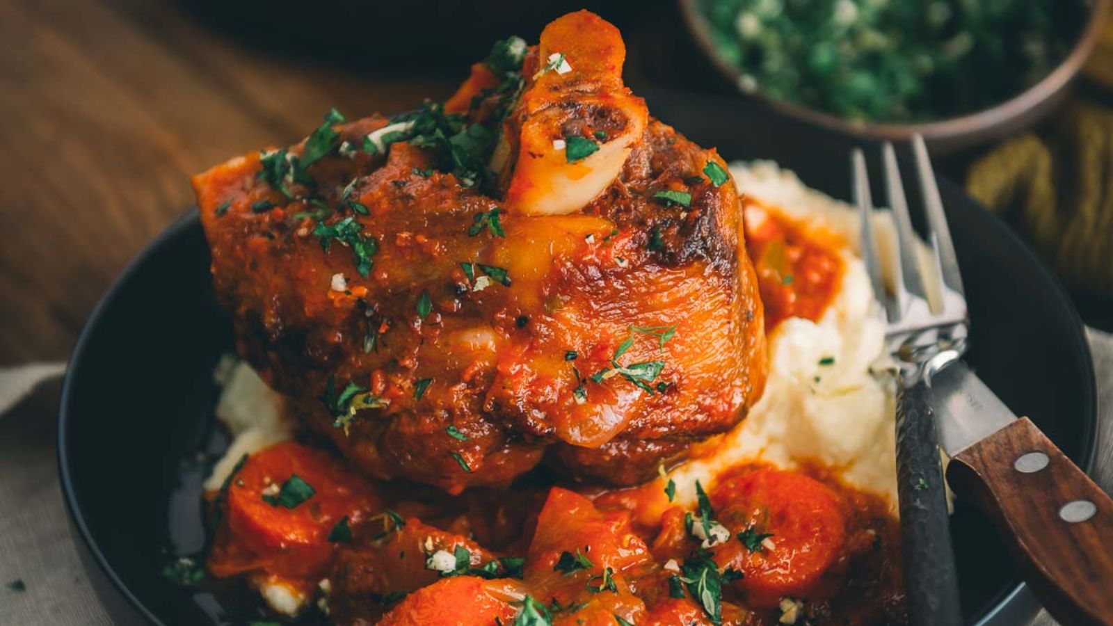 A plate of osso buco with braised meat, carrots, and sauce on mashed potatoes, garnished with chopped herbs, next to a fork.