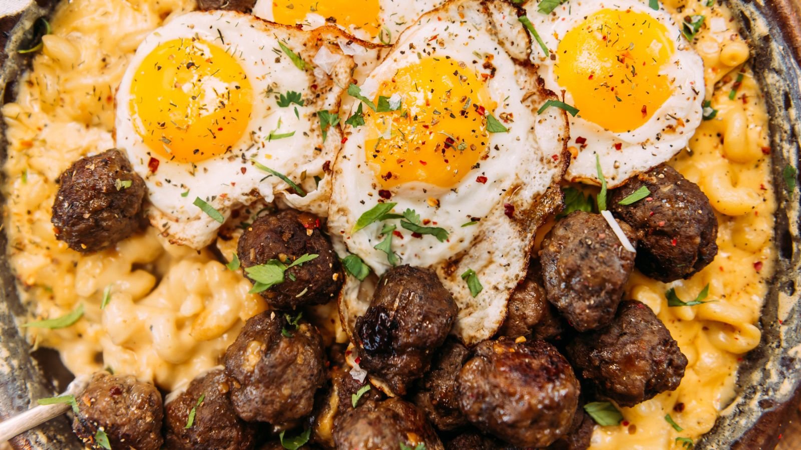 A dish of macaroni and cheese topped with three sunny-side-up eggs and seasoned meatballs, garnished with herbs.