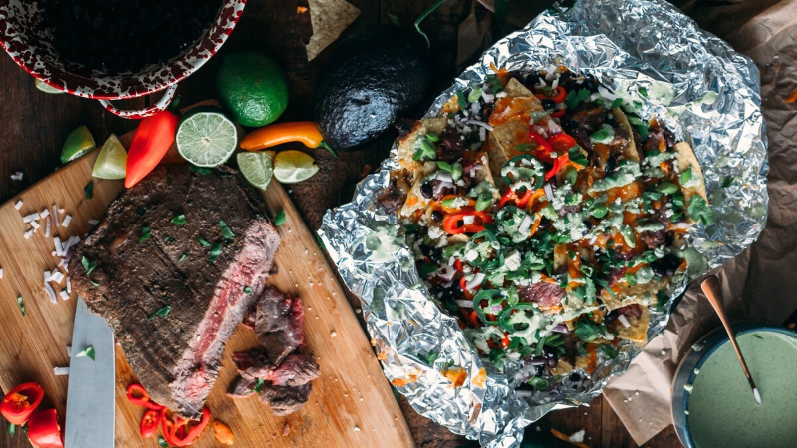 A foil tray with nachos topped with cheese, meat, and vegetables beside a cutting board with sliced steak and fresh peppers, lime, and avocado.