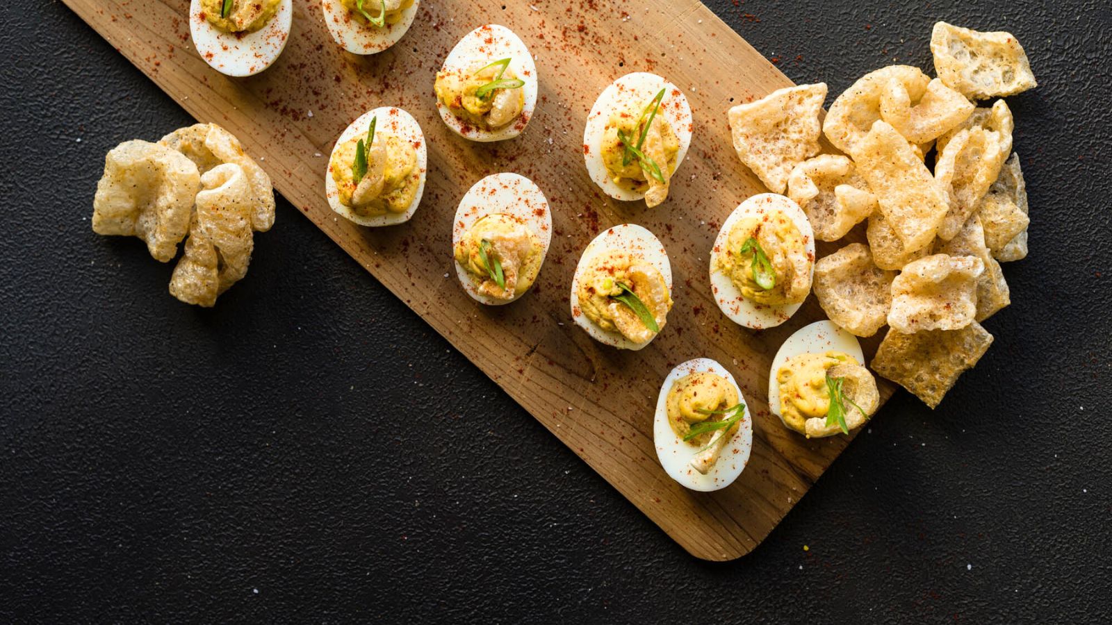 Deviled eggs on a wooden board garnished with spices and green herbs, accompanied by crispy pork rinds on a dark surface.