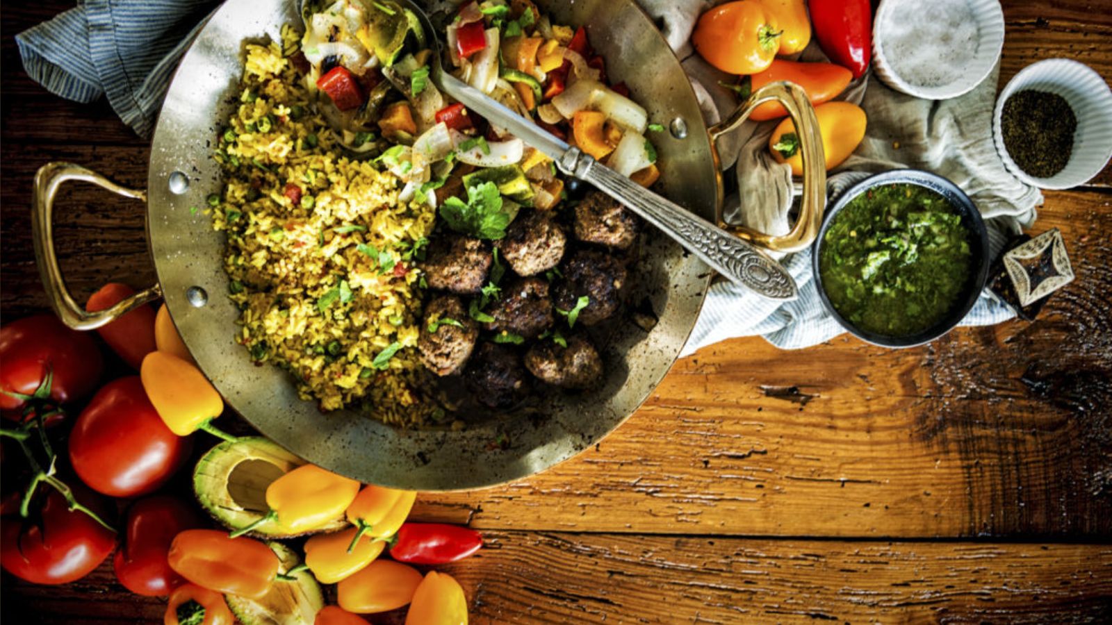 A brass pan with spiced rice, grilled meatballs, and vegetables. Beside it are colorful peppers, tomatoes, avocado, a small bowl of sauce, and seasoning on a wooden table.