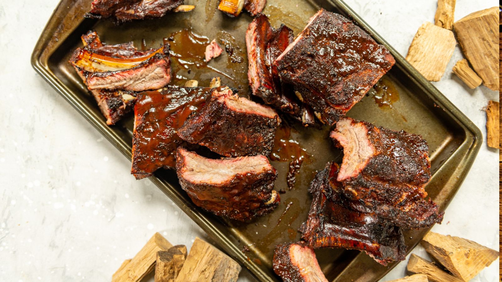 Barbecued beef ribs on a metal baking tray, surrounded by wood chips, against a light background.