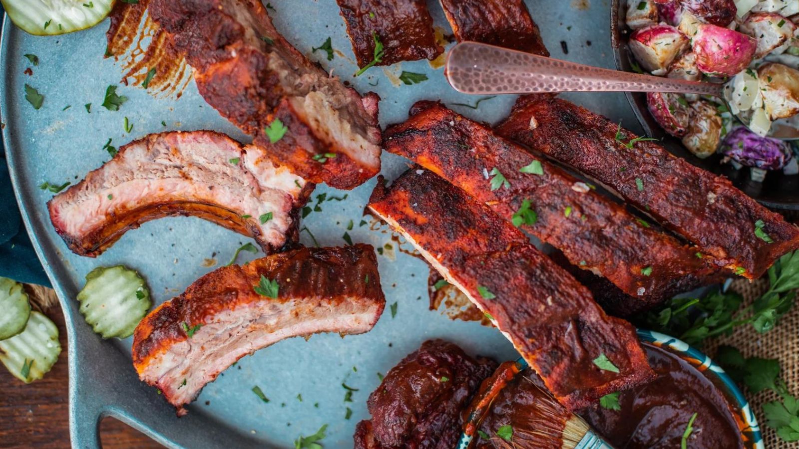 Plate of grilled ribs with parsley garnish, served with pickles and potato salad.