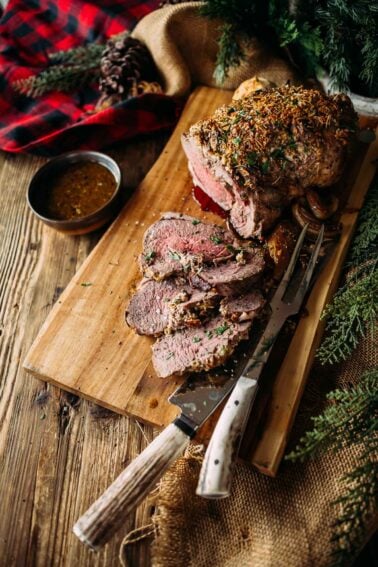 Sliced leg of lamb on a wooden board with herbs, a carving knife, fork, and a small bowl of sauce. Rustic setting with pine branches and a red plaid cloth in the background.