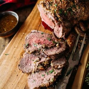 Slices of leg of lamb with herbs on a wooden board, accompanied by a carving knife and fork. A small bowl of gravy is nearby.