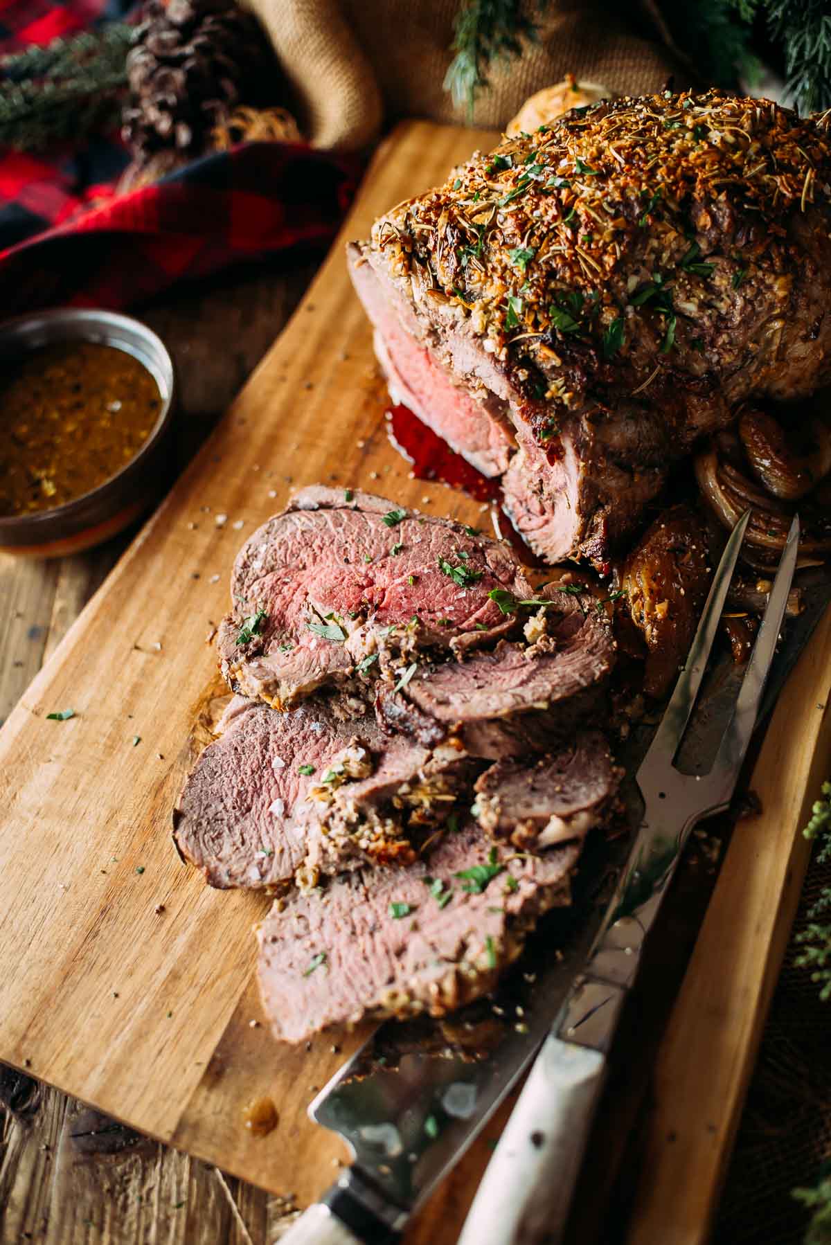 Slices of leg of lamb with herbs on a wooden board, accompanied by a carving knife and fork. A small bowl of gravy is nearby.