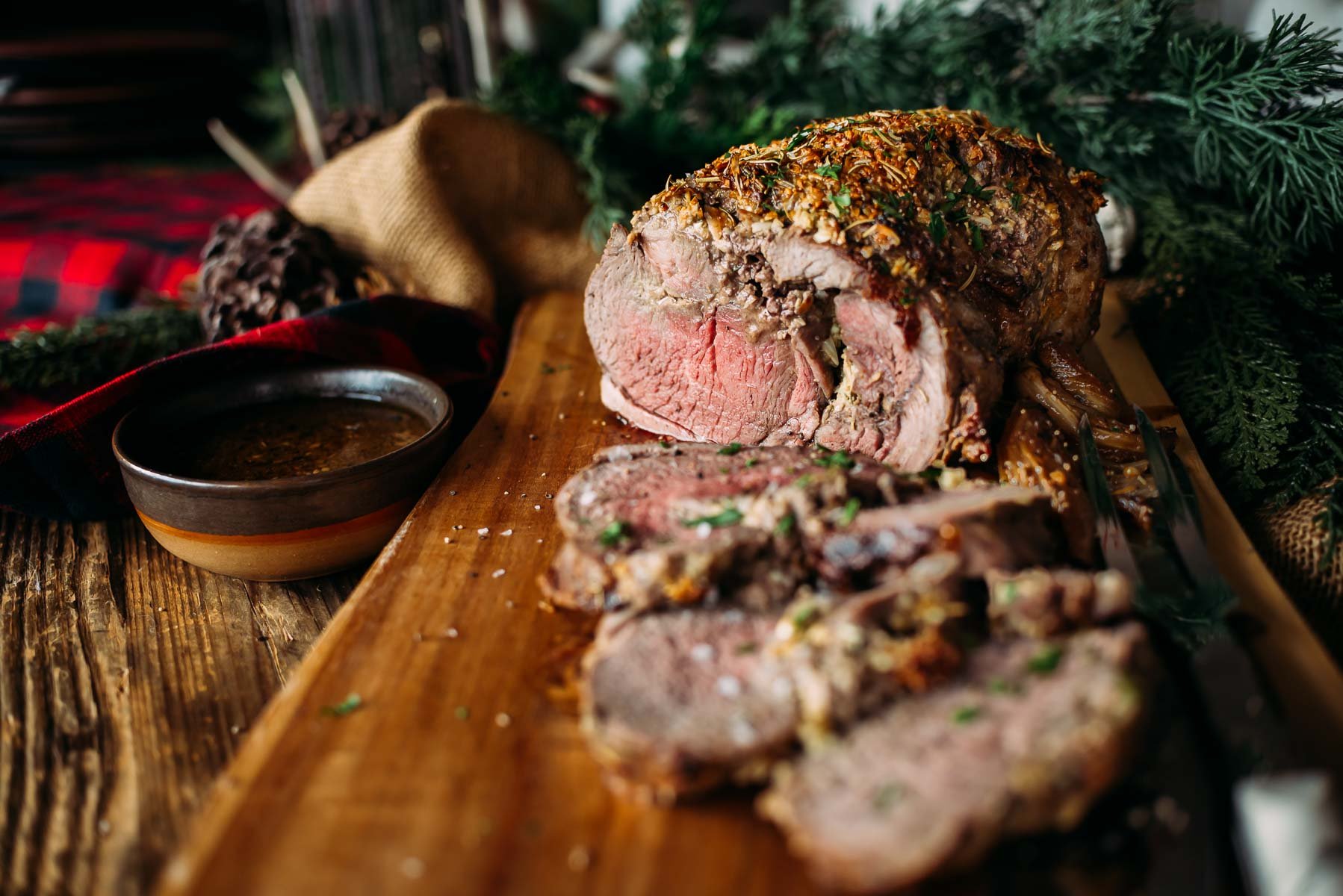 Sliced roasted leg of lamb on a wooden board with a bowl of sauce, garnished with herbs, set on a rustic table with holiday decorations.