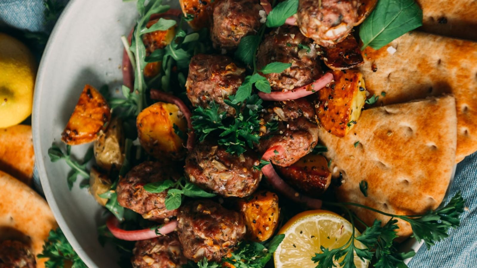 Plate of meatballs on a bed of greens with roasted potatoes, flatbread, lemon slice, and fresh herbs.