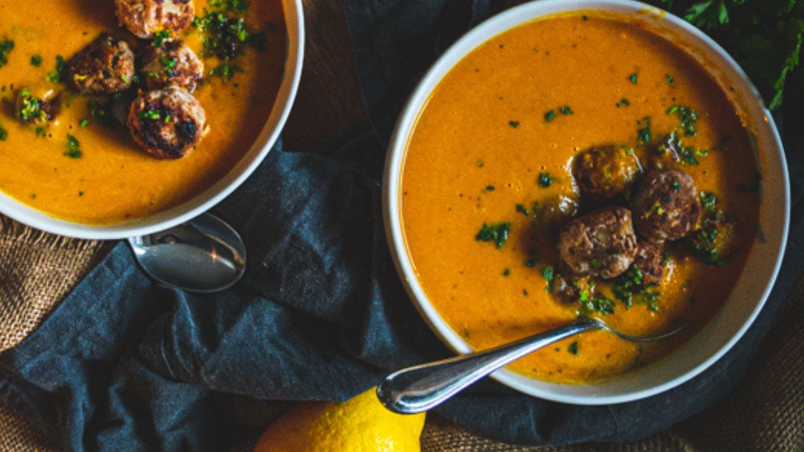 Two bowls of orange soup with meatballs and herbs, accompanied by a lemon, on a dark cloth background.