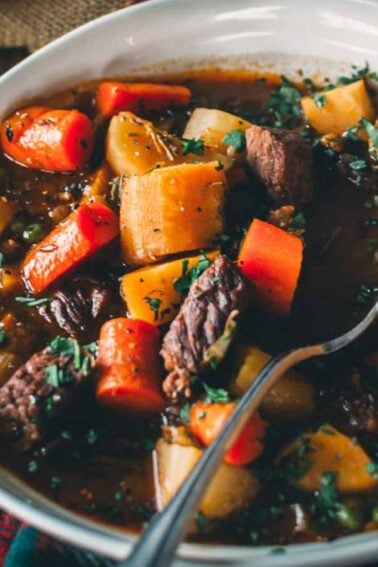 A bowl of beef stew with chunks of meat, carrots, and potatoes, garnished with herbs. A spoon rests in the bowl, and a slice of toasted bread is placed on the rim.