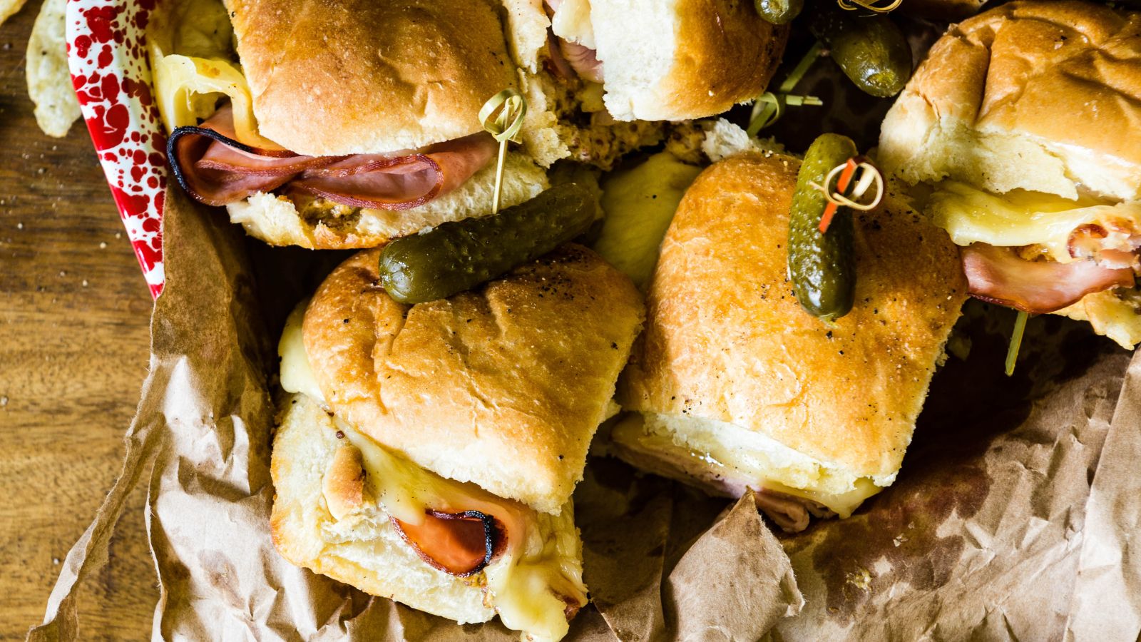 Close-up of ham and cheese sliders on crumpled parchment paper, each topped with a pickle and secured with a toothpick.