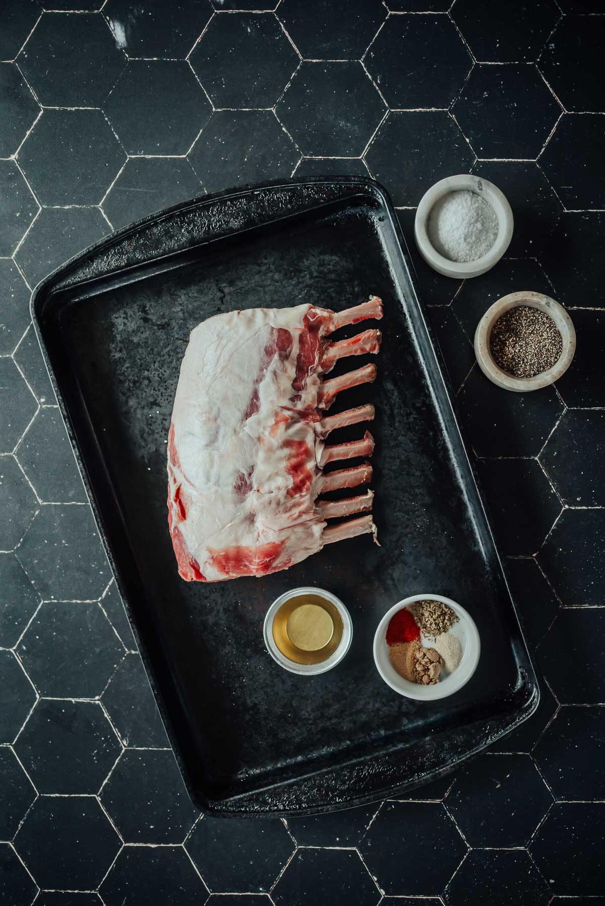 Raw rack of boar on a baking tray, surrounded by small bowls of spices and seasoning, on a dark hexagonal tiled surface.