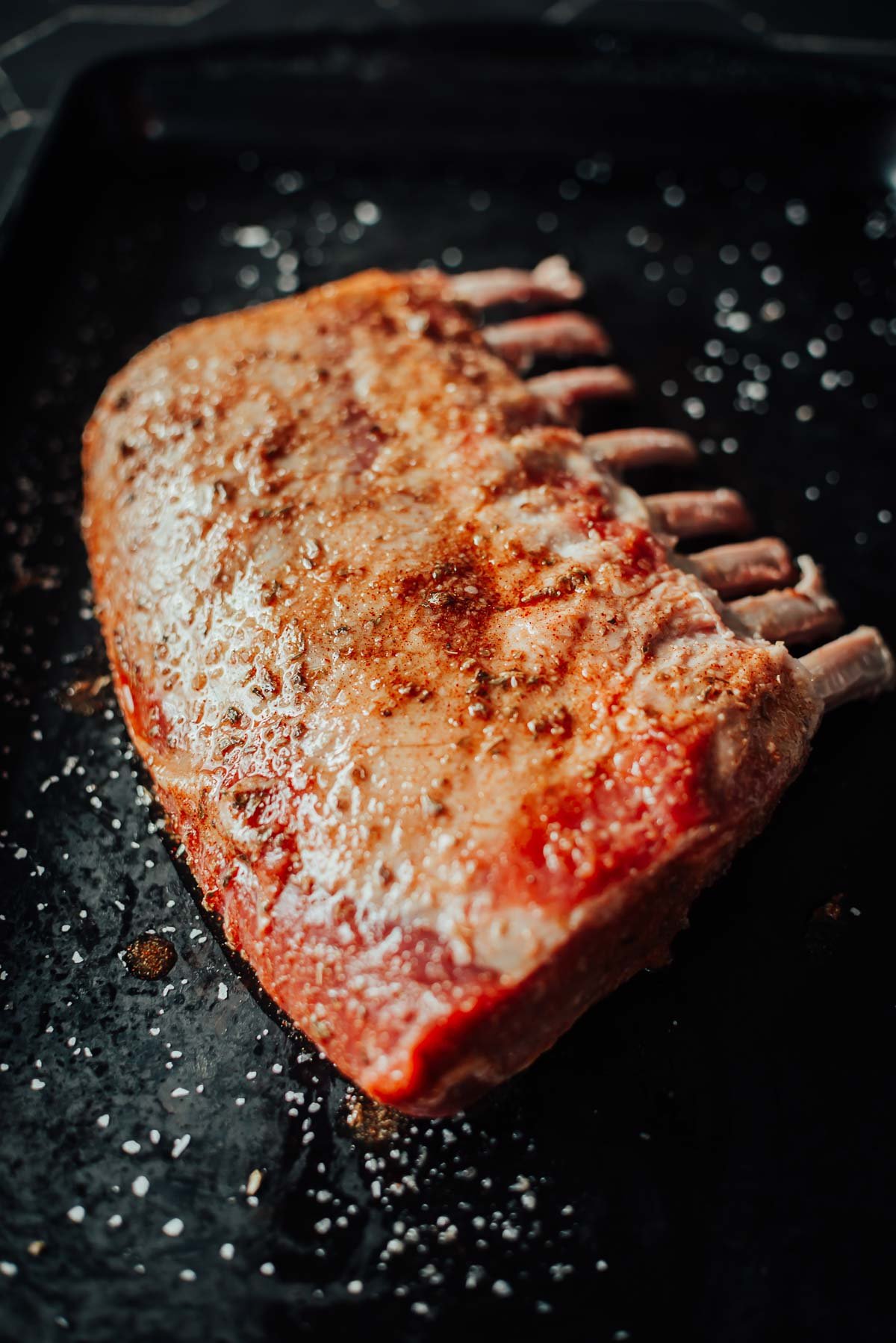 A seasoned rack of boar on a dark baking tray sprinkled with salt.