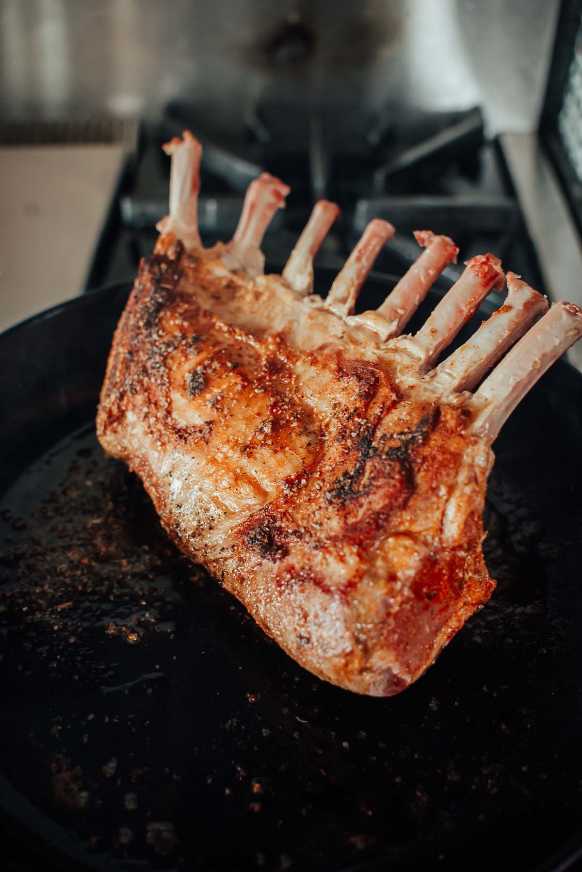 A rack of roasted boar with a crispy brown crust sits in a black pan on a stovetop.