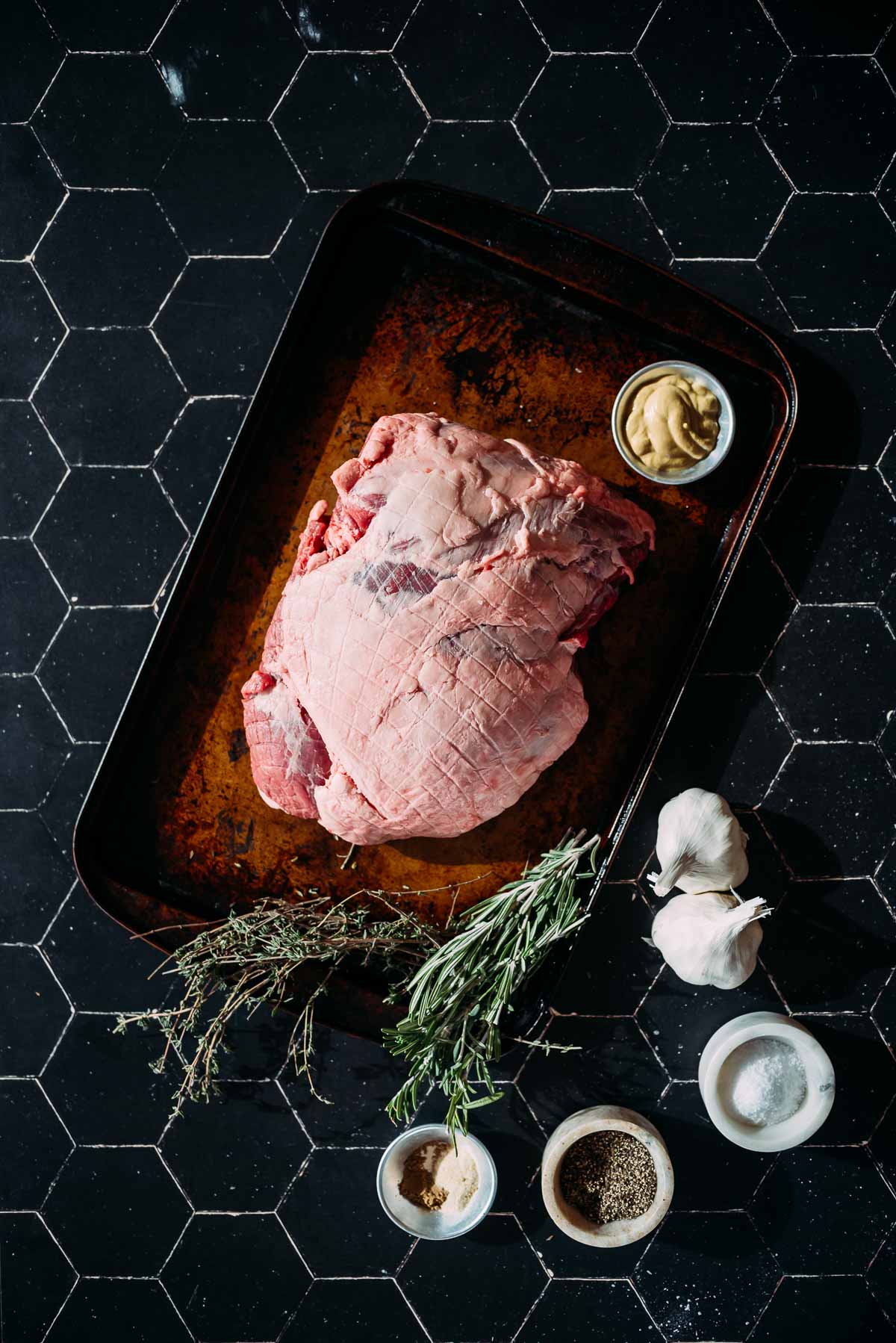 A raw boneless leg of lamb on a baking tray, surrounded by bowls of mustard, salt, and pepper, with fresh thyme, rosemary, and garlic on a black tiled surface.