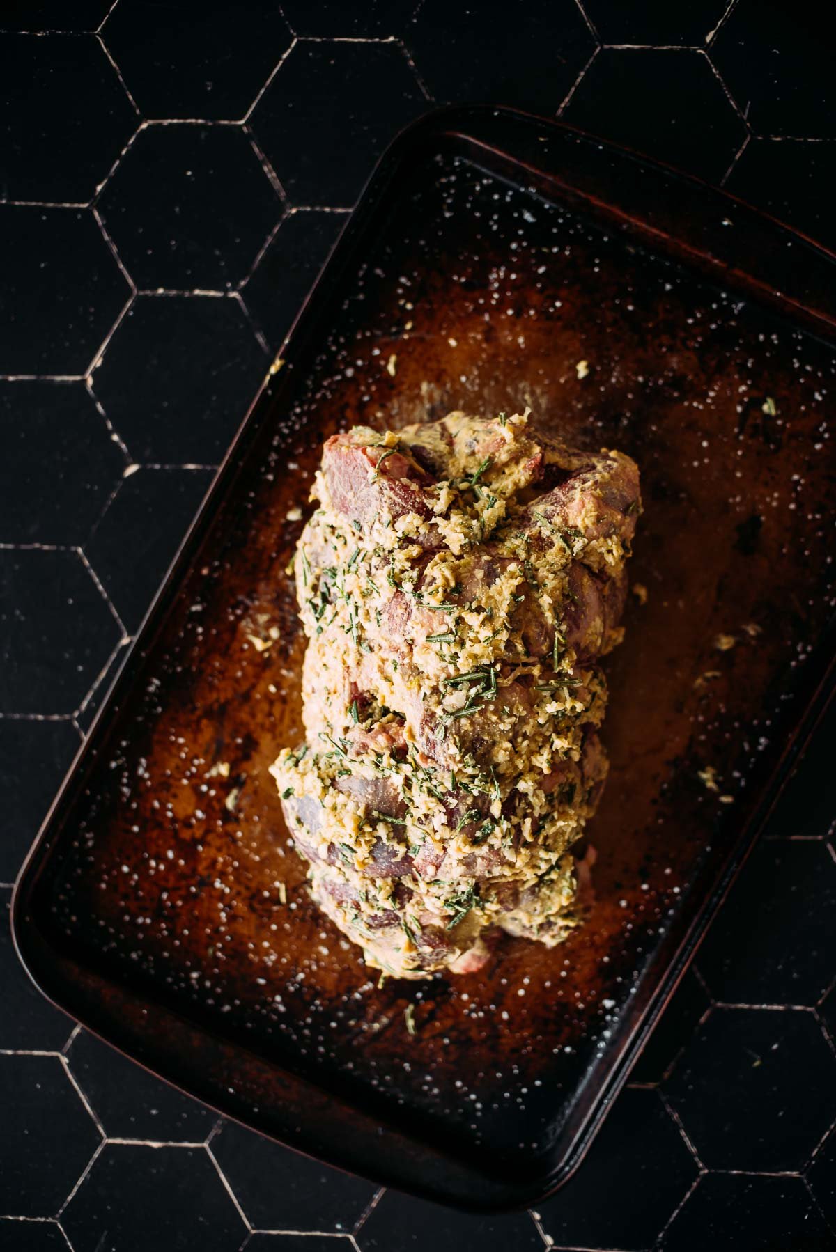 Leg of lamb covered in mustard and herbs and seasoning on a dark baking tray.