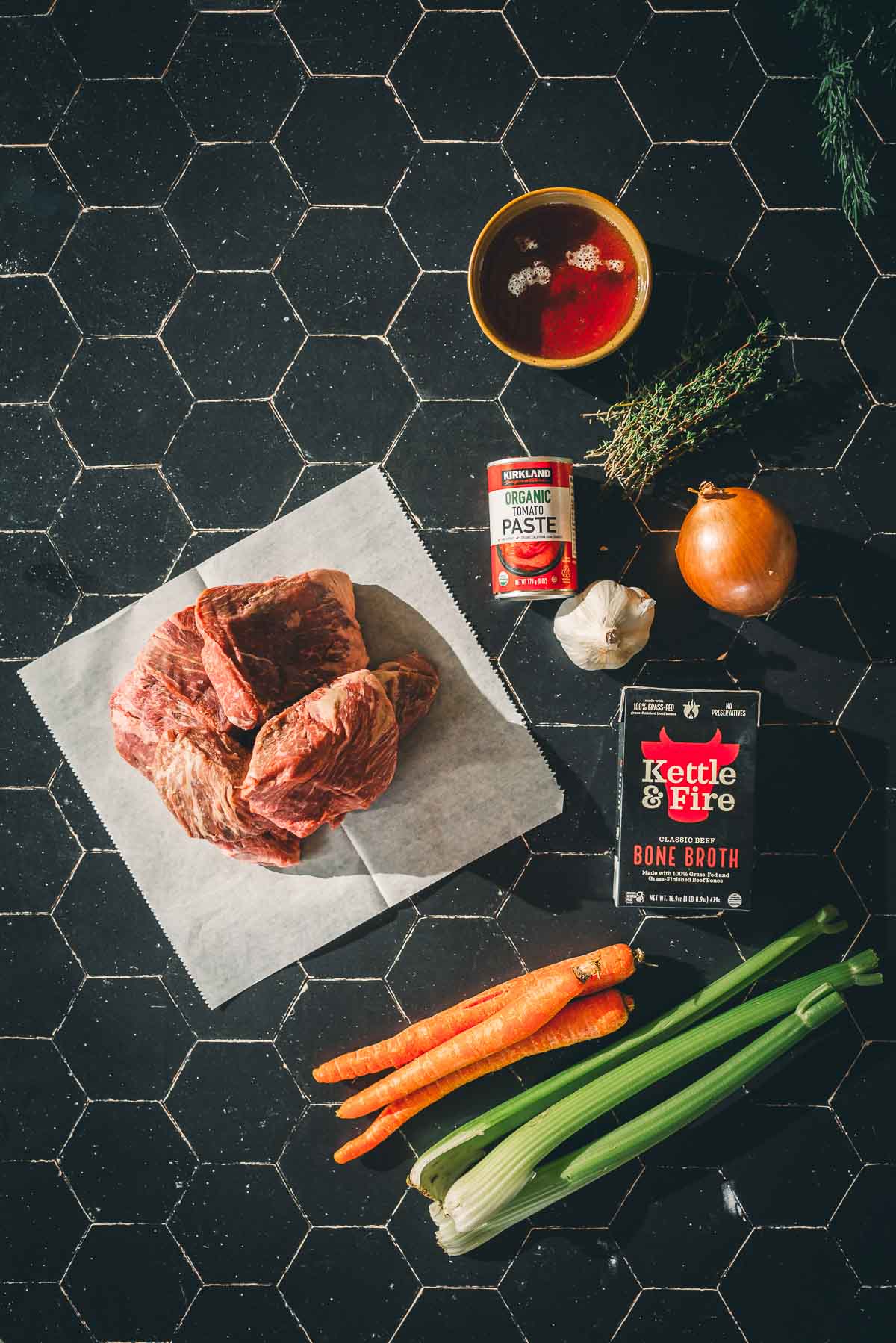 Ingredients for sous vide short ribs on a dark tile surface: raw beef on paper, tomato paste, bone broth, garlic, onion, carrots, celery, thyme, and a bowl of red liquid.