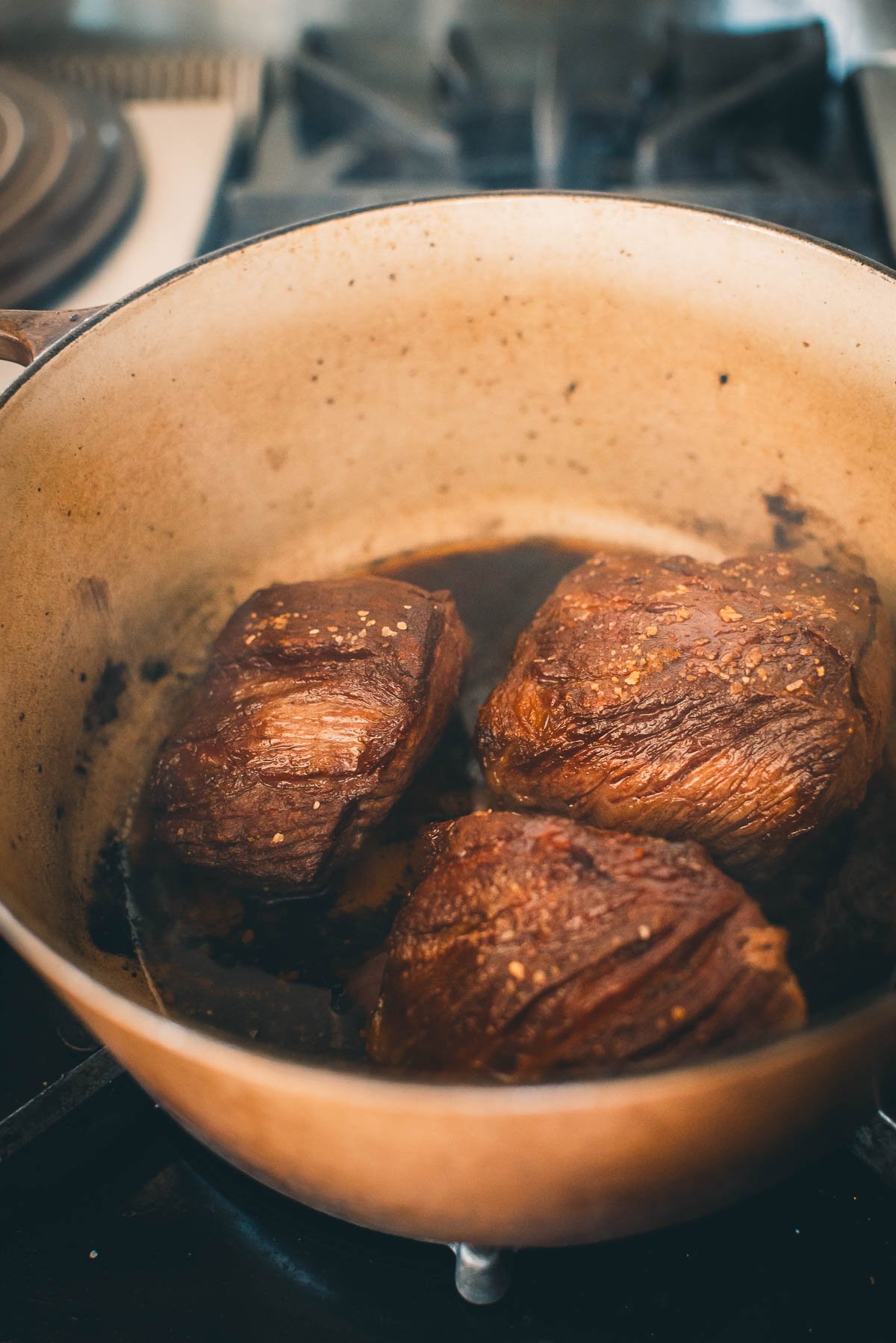 Three pieces of browned short ribs cooking in a pot on a stovetop.