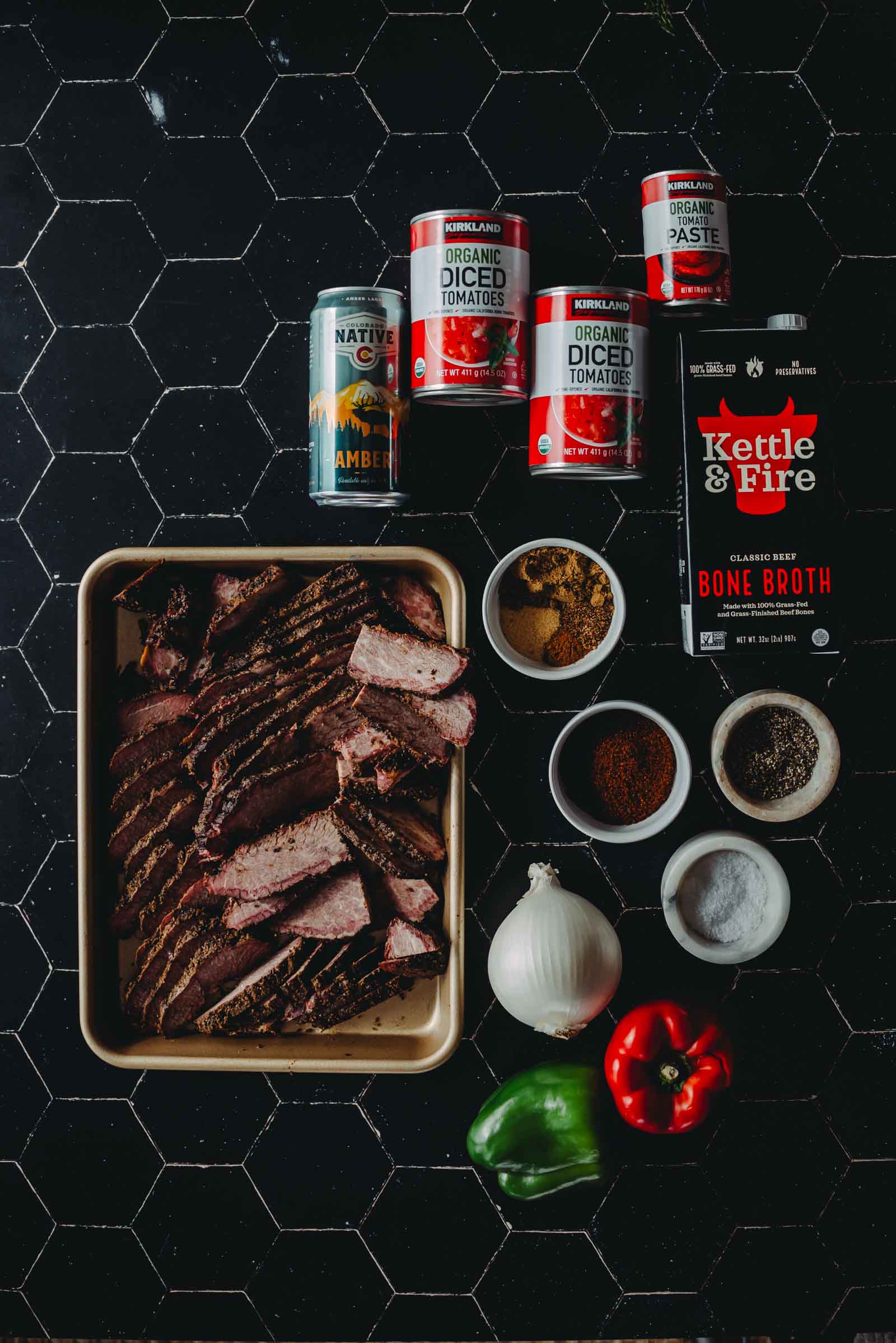 Sliced smoked brisket on a tray, surrounded by cans of tomatoes, tomato paste, bone broth, whole onion, green and red bell peppers, and small bowls of spices on a dark tile surface.