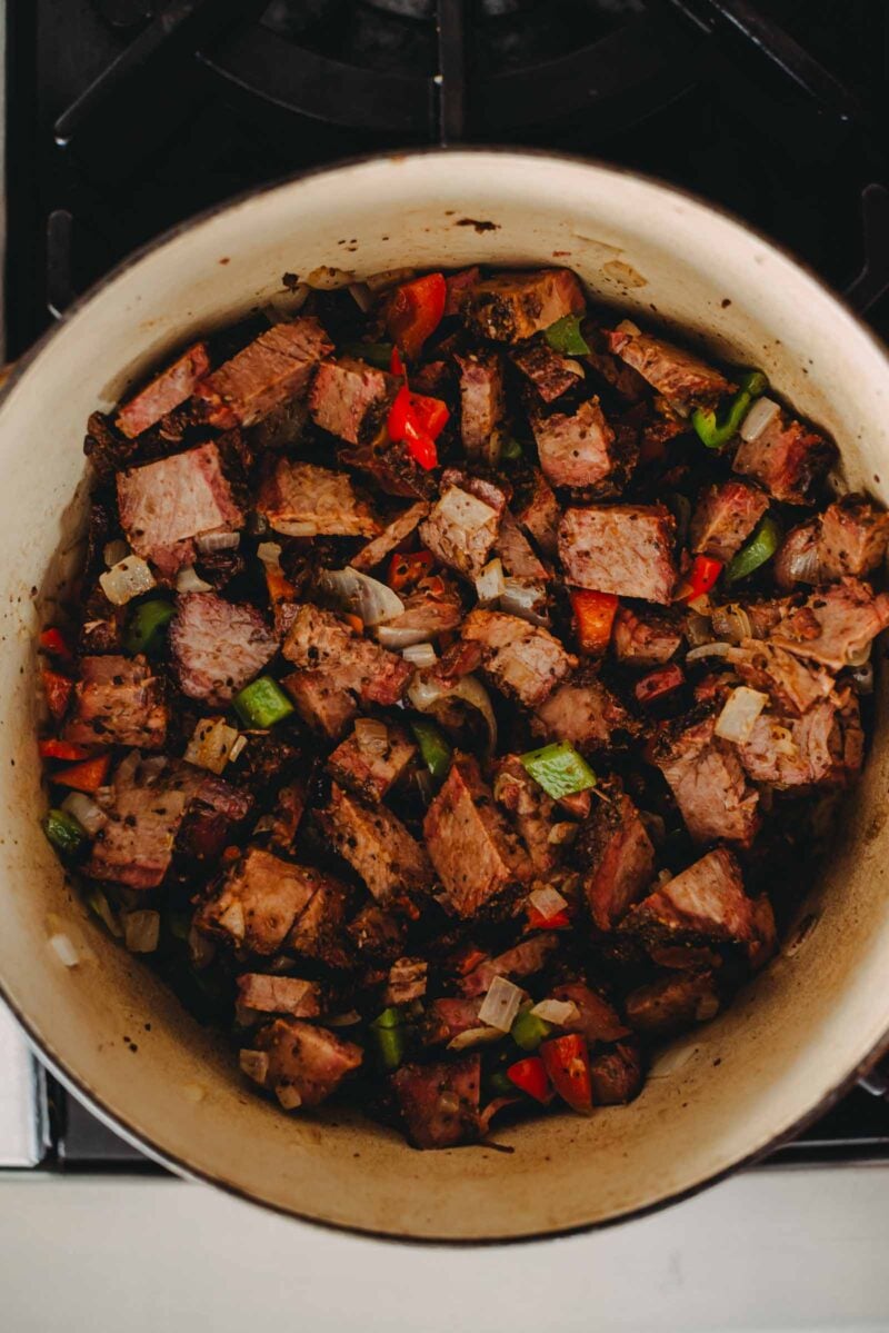 Chunks of smoked beef brisket with red and green bell peppers and onions in a pot on a stove.