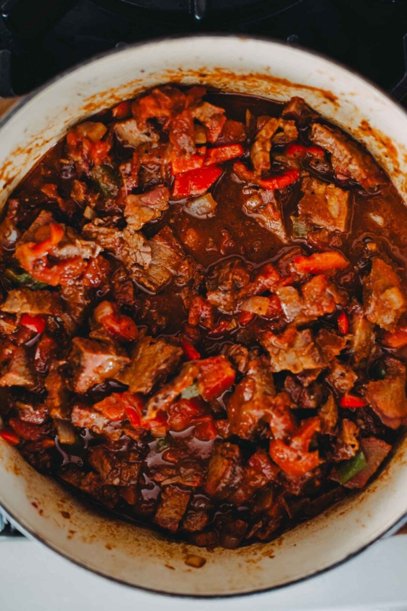 A pot filled with a brisket chili containing chunks of meat, red bell peppers, and a rich, dark sauce.