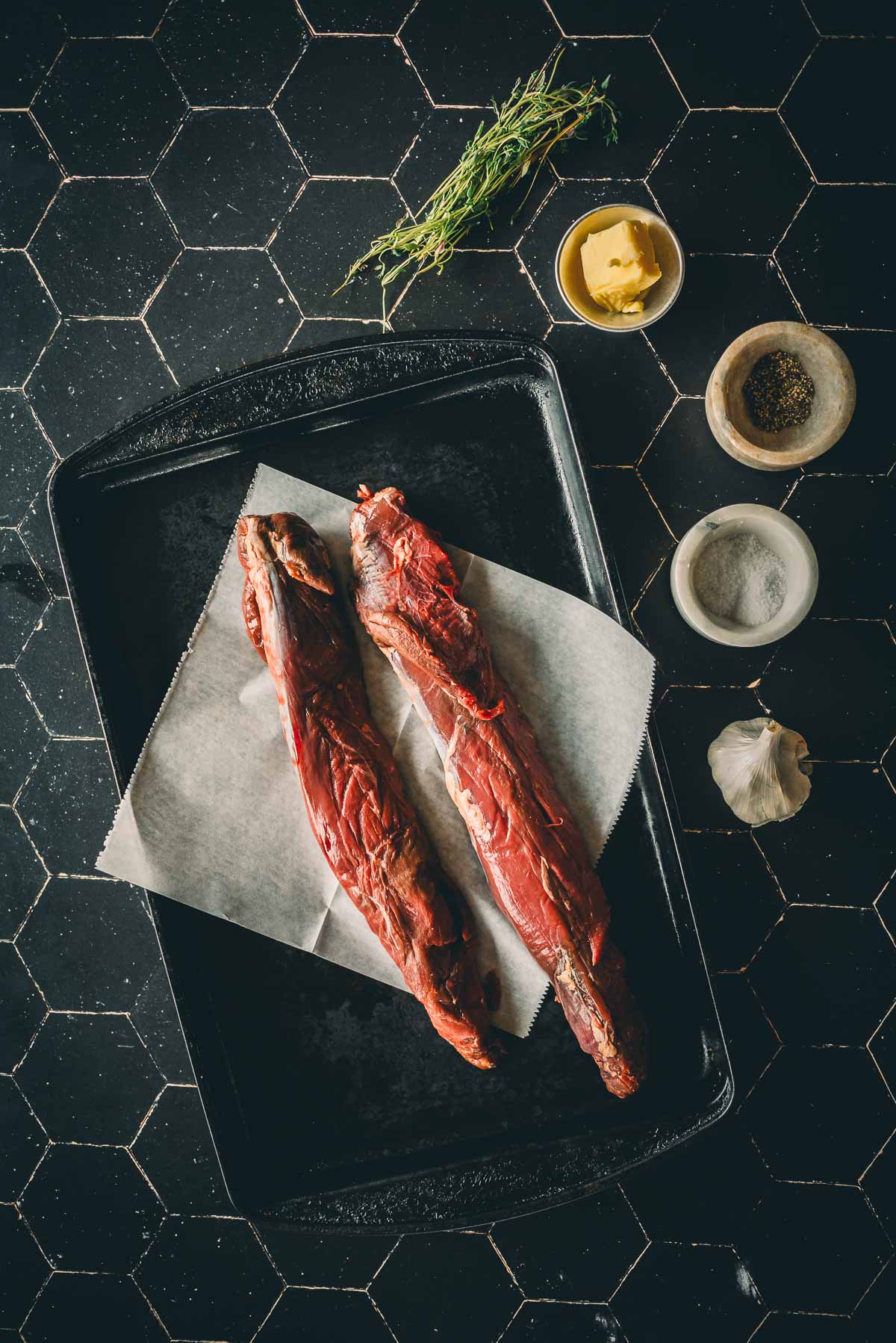 Two raw venison tenderloins on a baking tray with parchment paper, surrounded by thyme, butter, salt, pepper, and garlic on a black hexagonal tile surface.