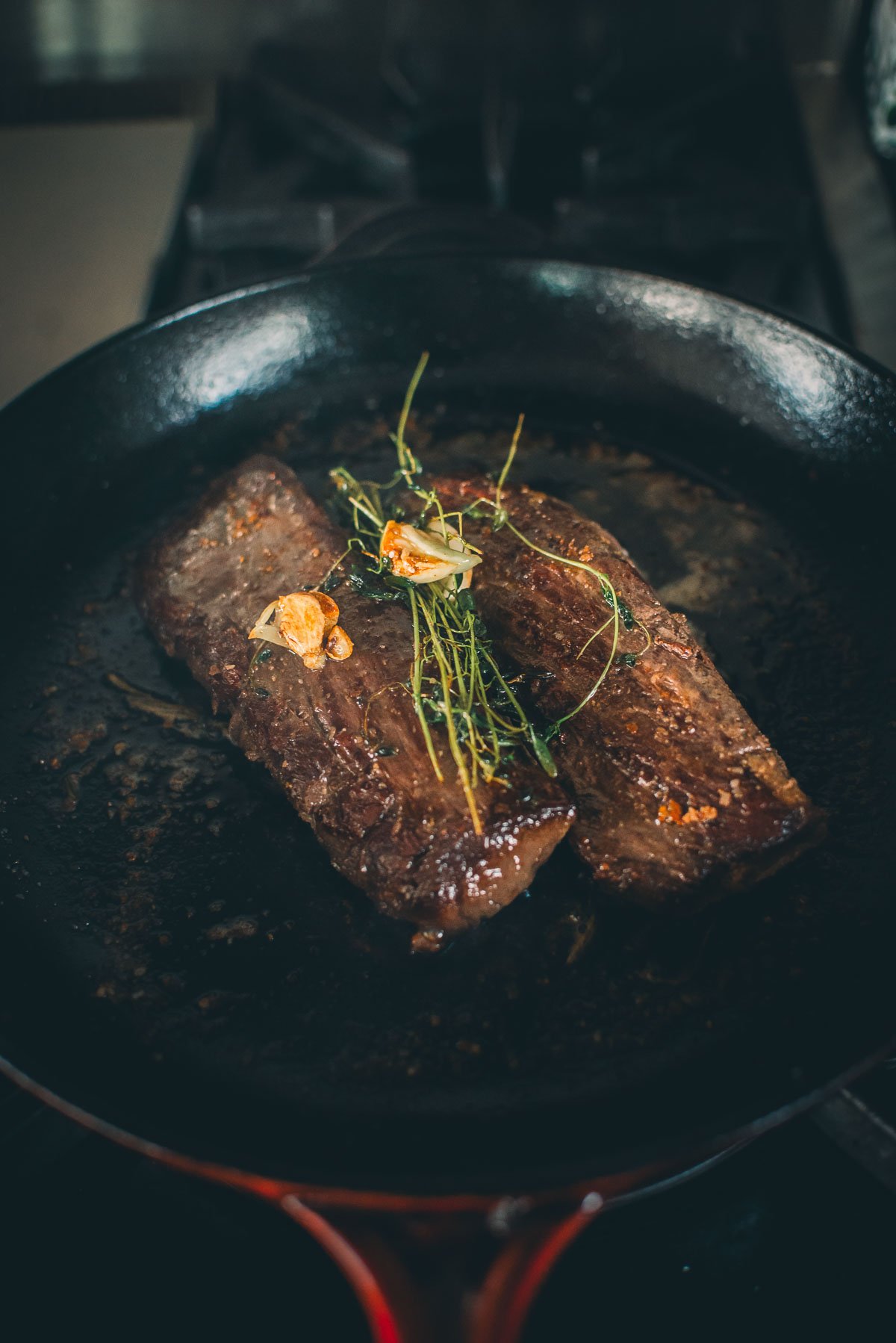 Two venison tenderloins are cooking in a pan, topped with herbs and garlic cloves.