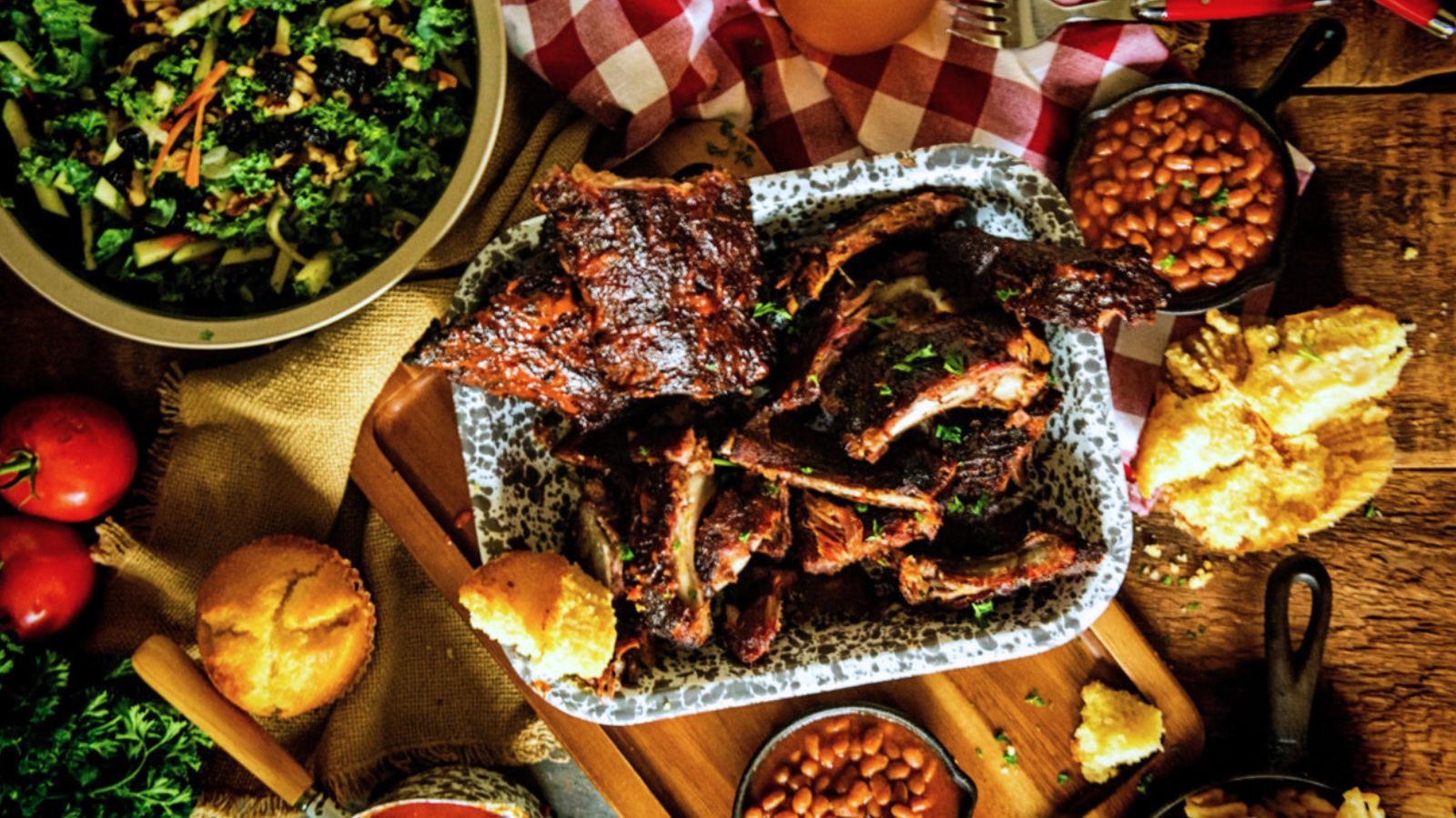 A rustic wooden table set for a meal with barbecue ribs on a platter, a bowl of salad, baked beans, cornbread muffins, and tomatoes, on a checkered cloth.