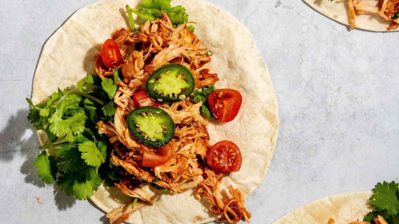 A tortilla with shredded chicken, sliced jalapeños, cherry tomatoes, and cilantro on top, placed on a light surface.