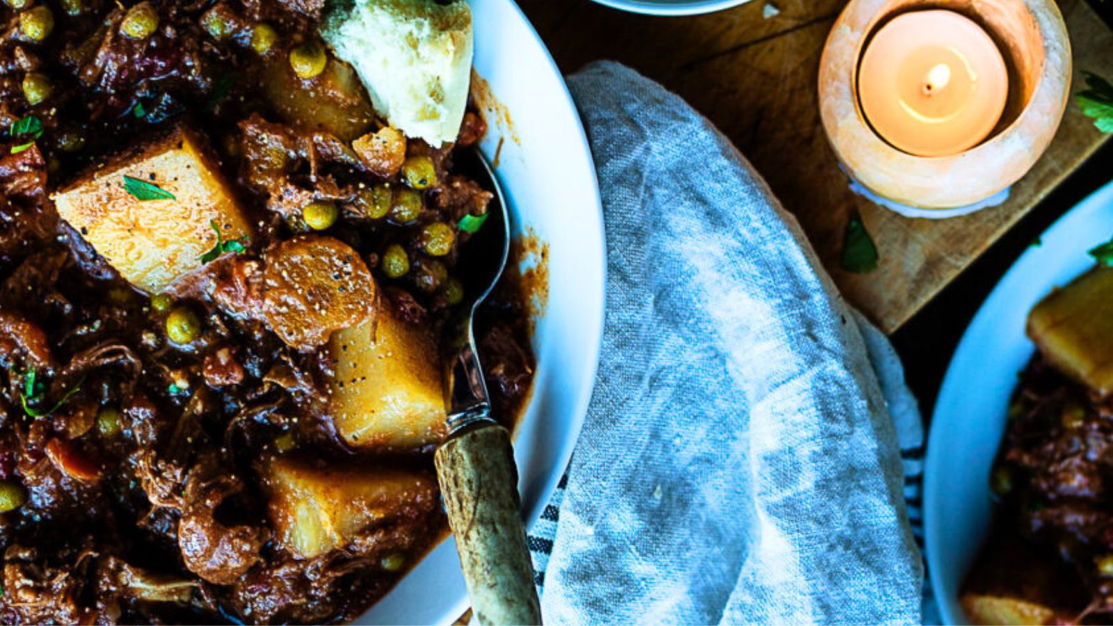 A bowl of thick stew with potatoes, peas, and meat. A spoon rests inside, next to a lit candle and a napkin.