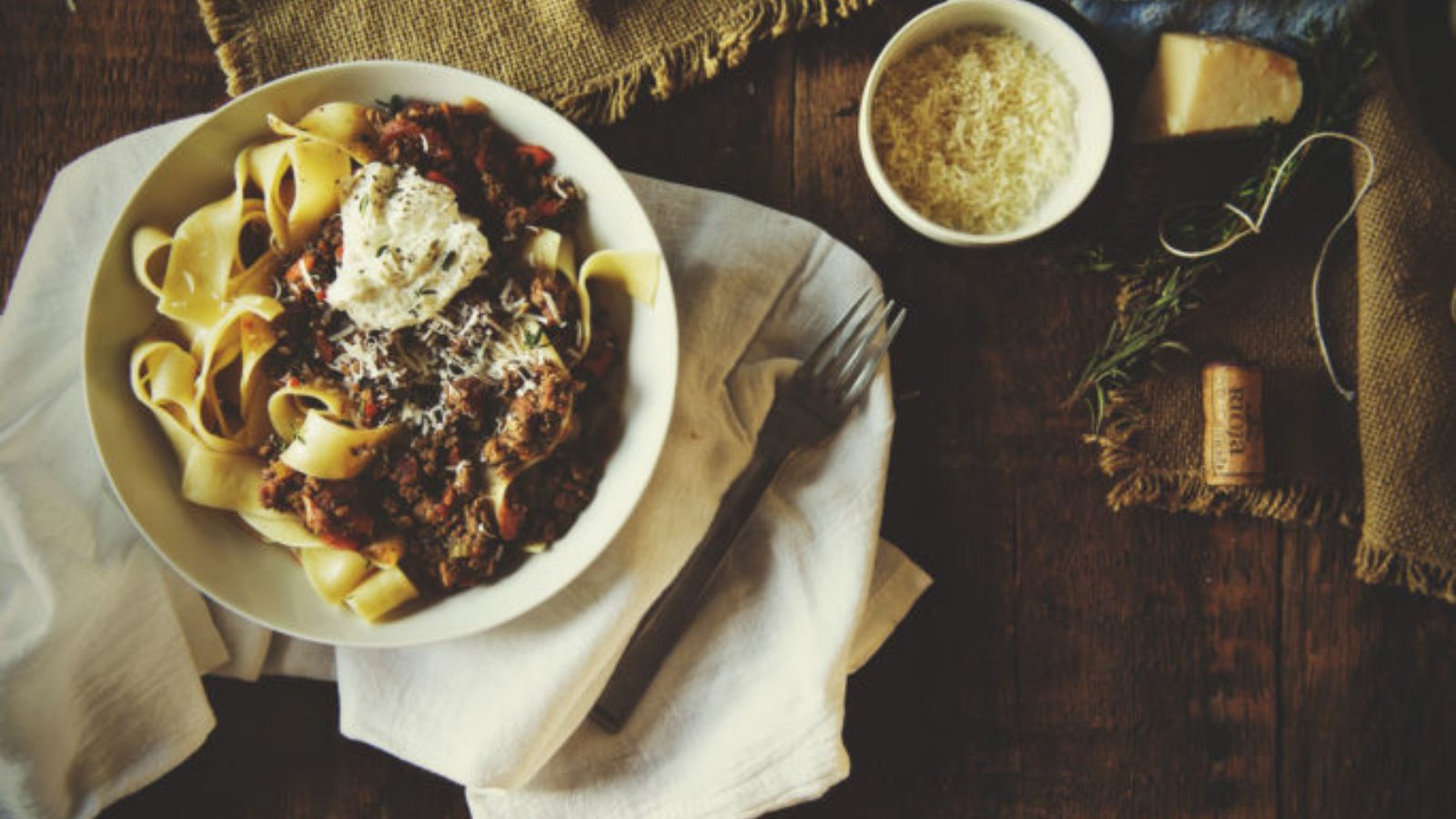 A plate of pasta with meat sauce and ricotta, on a wooden table with a fork, small bowl of grated cheese, a block of cheese, and a cork.