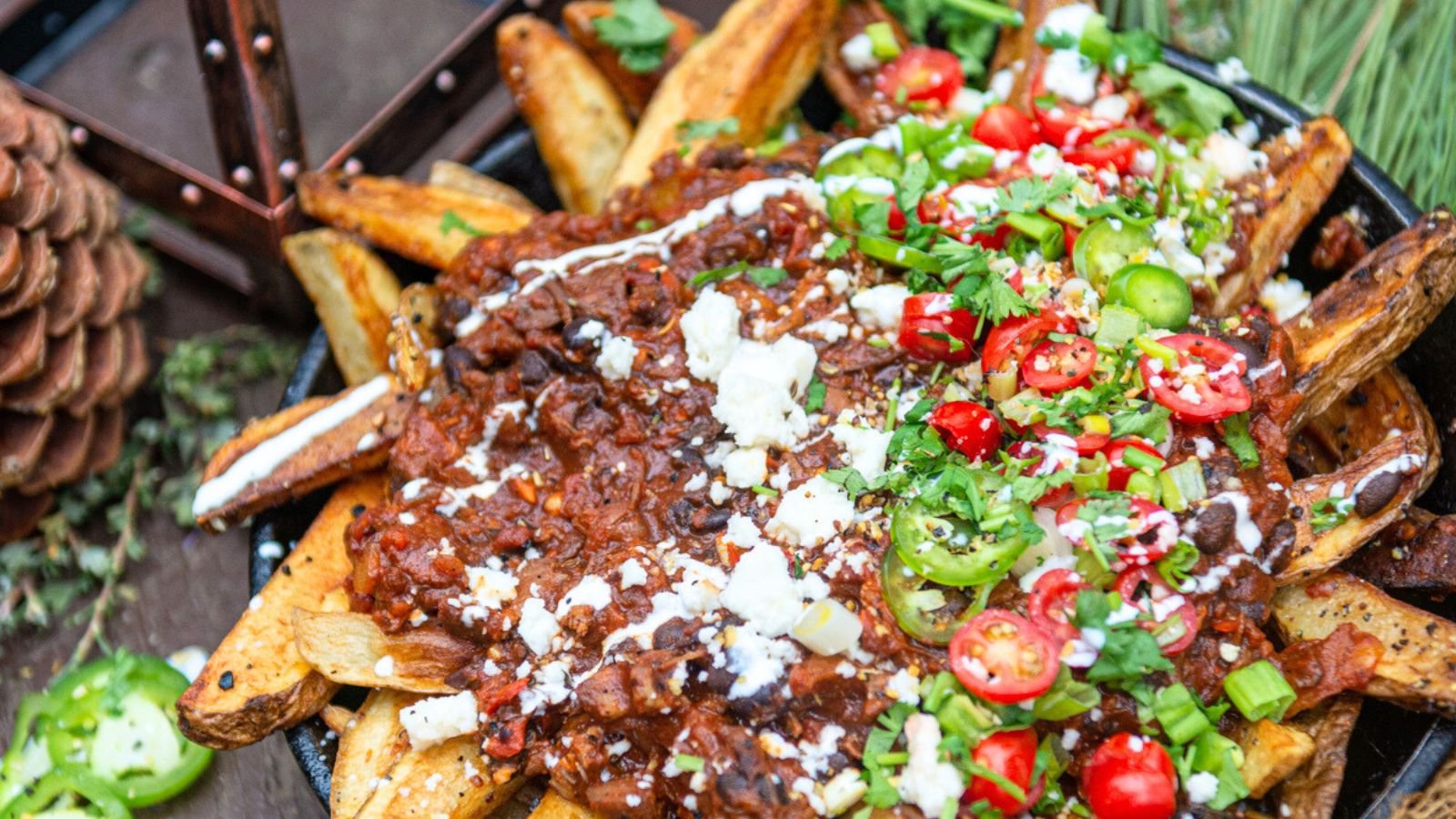 Loaded fries inspired by the best chili recipes, topped with diced tomatoes, jalapeños, cheese, sour cream, and chopped herbs on a rustic wooden table.