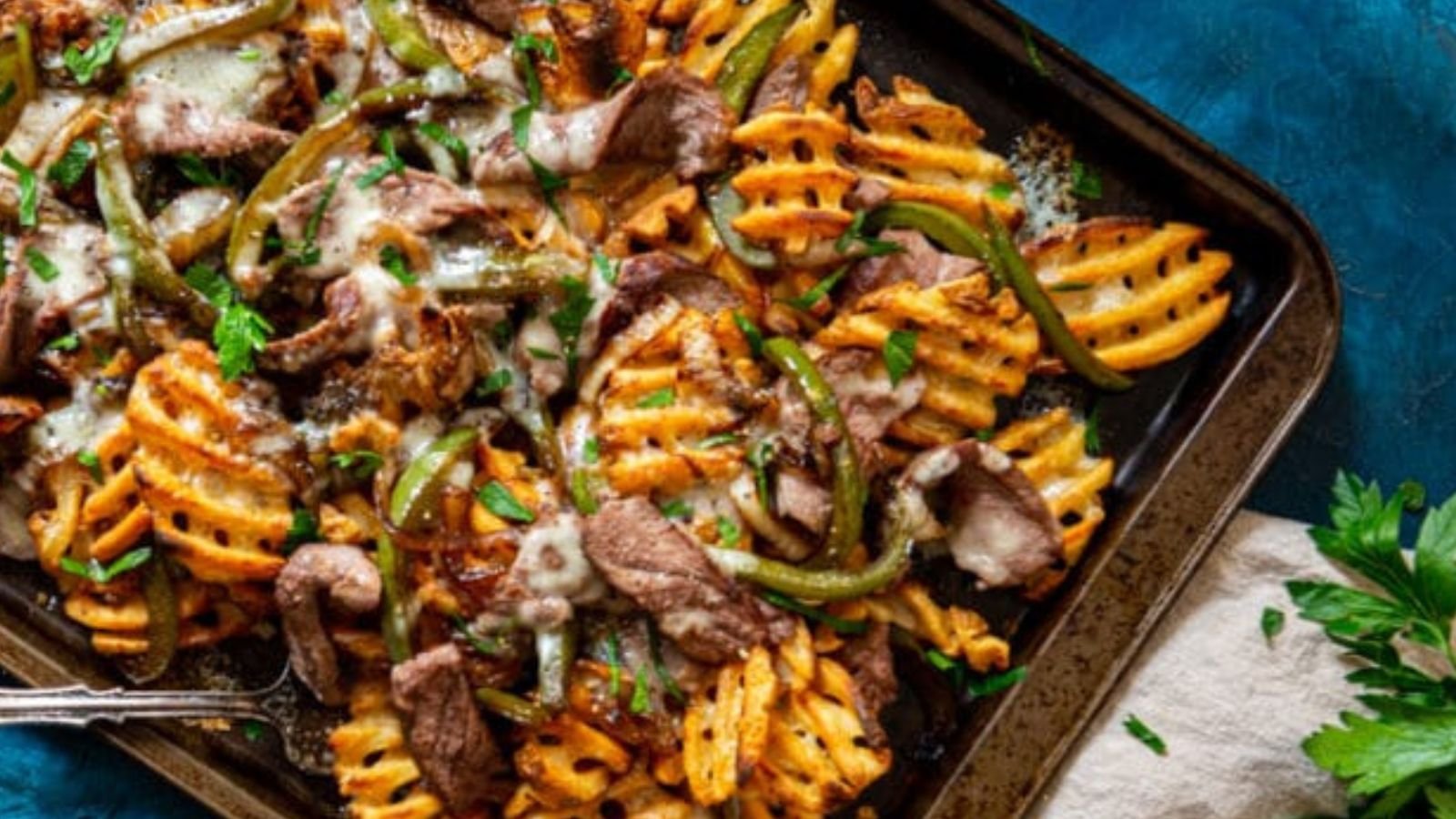 A tray of waffle fries topped with sliced beef, green peppers, and melted cheese. Parsley is scattered on top.