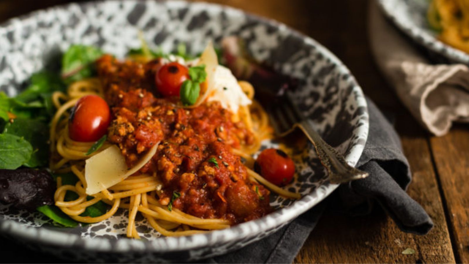 A plate of spaghetti with tomato-based sauce, cherry tomatoes, and greens, topped with cheese slices. A fork rests on the side.