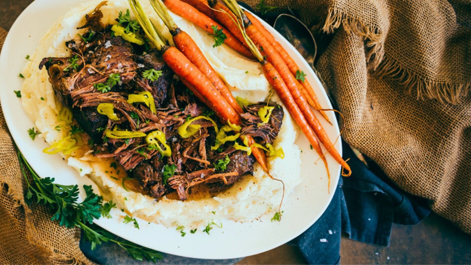 A plate with shredded roast beef, mashed potatoes, and whole carrots garnished with herbs and sliced peppers.