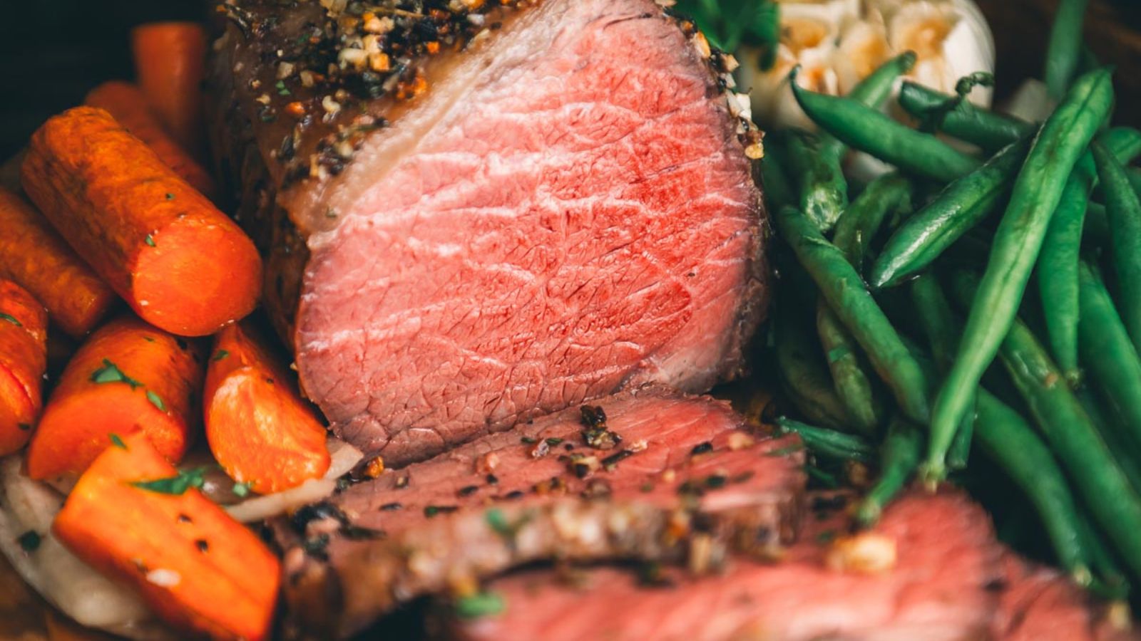 Sliced roast beef with seasonings, surrounded by green beans, carrots, and garlic cloves.