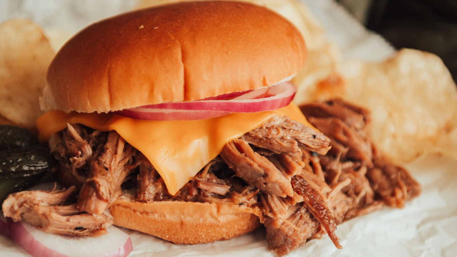 Pulled pork sandwich topped with cheese and red onions on a bun, served with pickles and potato chips.