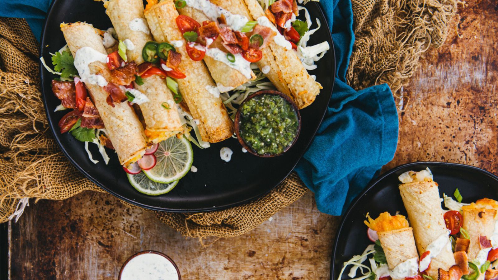 A plate of rolled tacos with toppings like tomatoes and jalapeños, served with green sauce on a distressed wooden table.