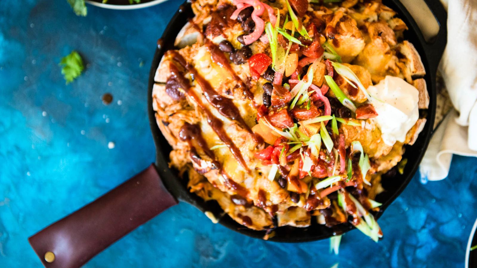 A skillet filled with loaded nachos topped with cheese, sour cream, sliced green onions, and diced tomatoes on a blue background.