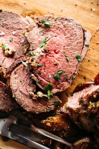Sliced leg of lamb with herbs on a wooden cutting board, accompanied by a carving knife and fork.