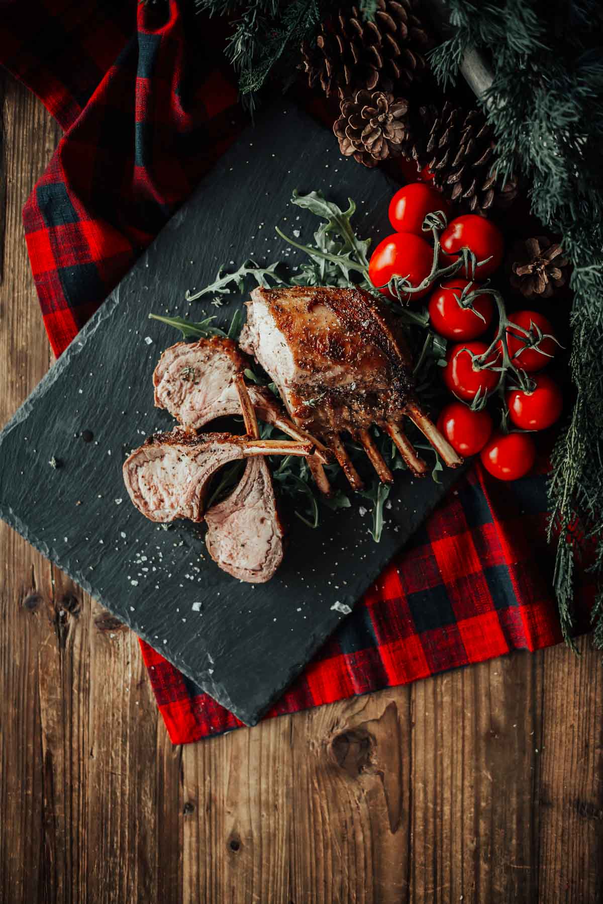 Rack of boar with sliced portions on a slate board, garnished with arugula and accompanied by a bunch of cherry tomatoes. Set on a plaid cloth over a wooden table.