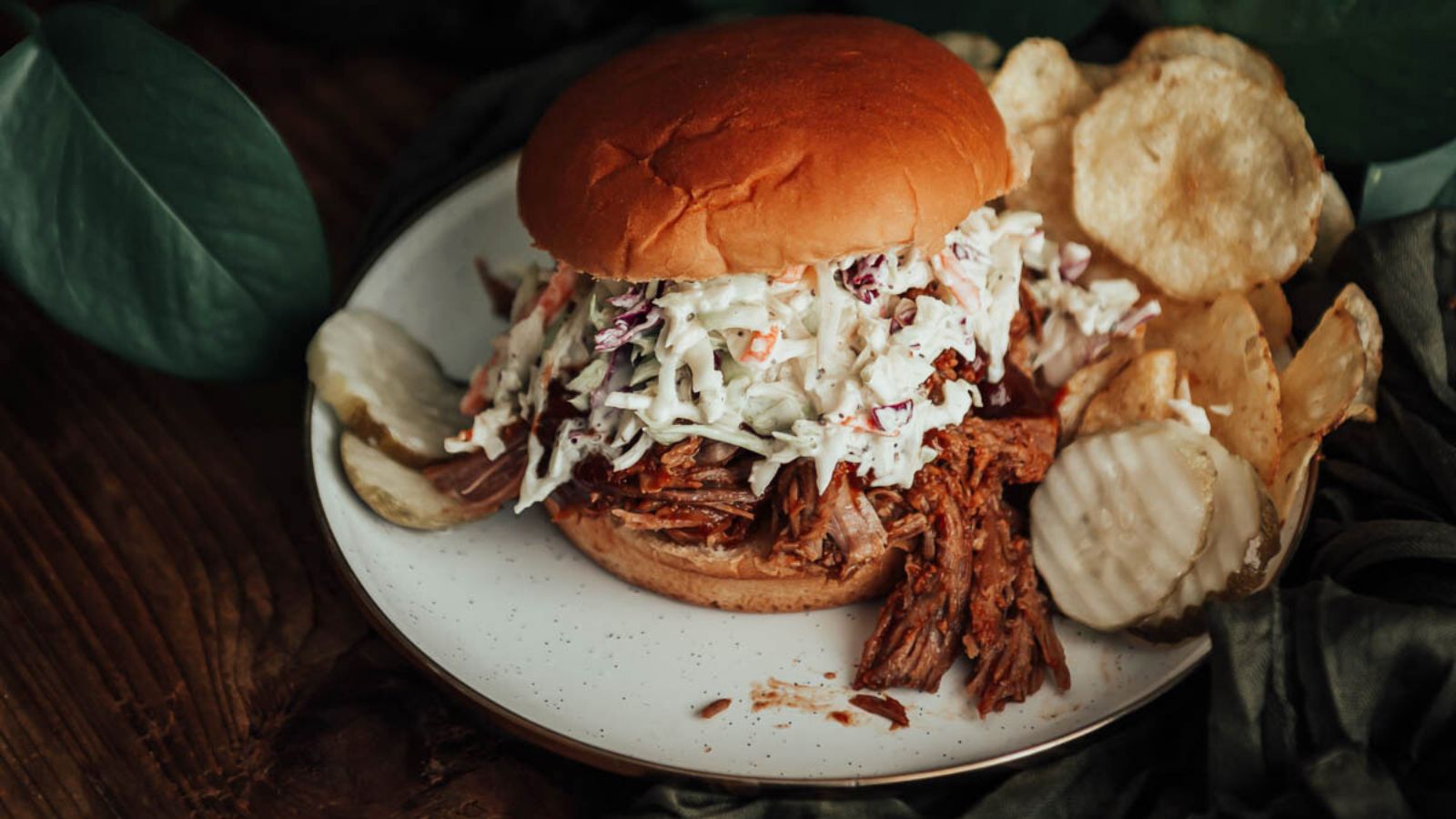 Pulled pork sandwich with coleslaw on a bun, served on a white plate with potato chips and a pickle slice, set against a wooden table with green leaves in the background.