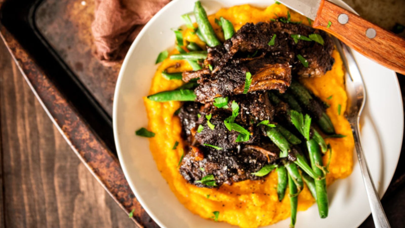 A plate of braised short ribs on a bed of mashed sweet potatoes with green beans, garnished with chopped parsley and served with a knife.