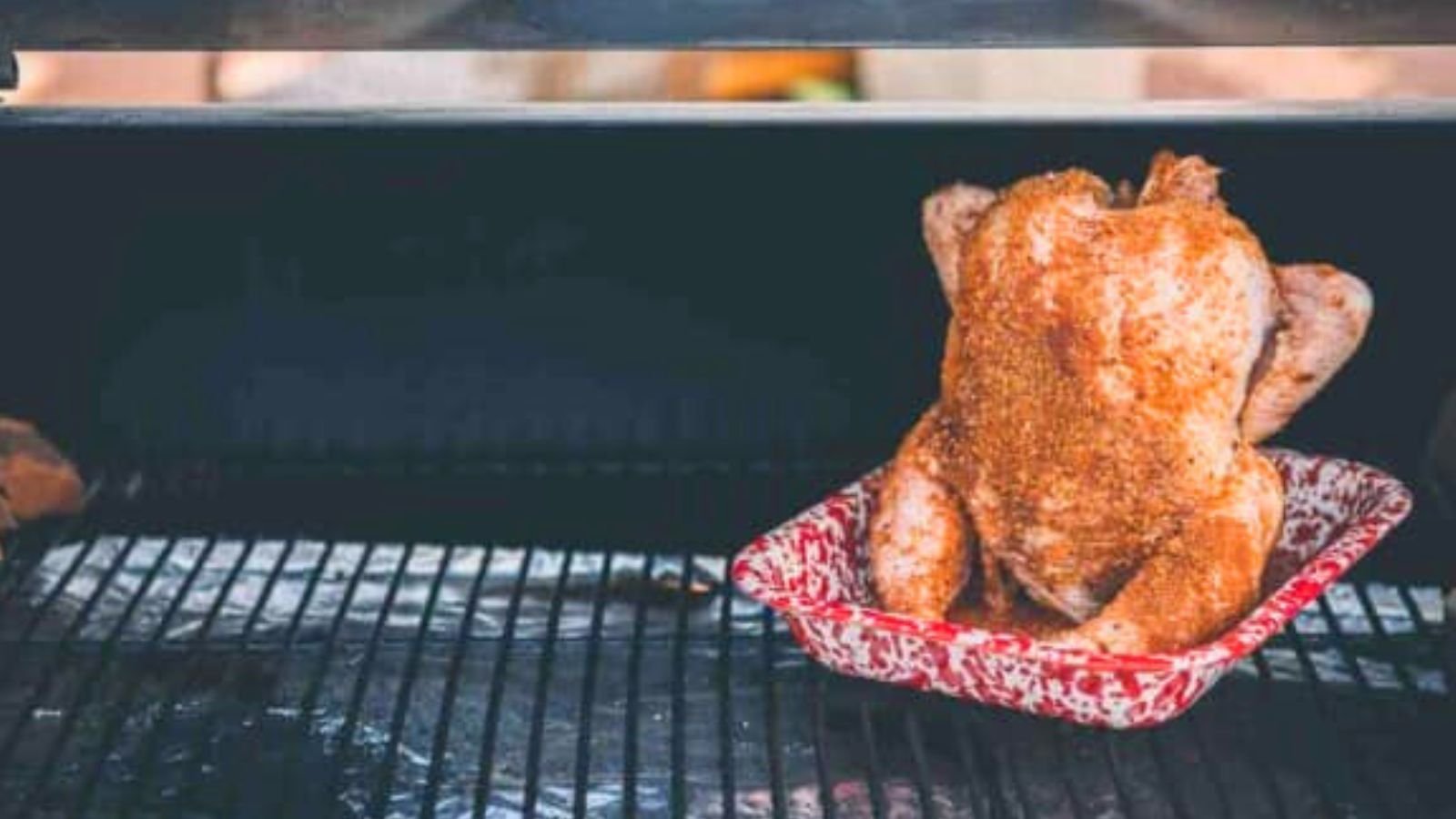 Whole chicken seasoned and placed upright in a red speckled roasting pan on a grill.