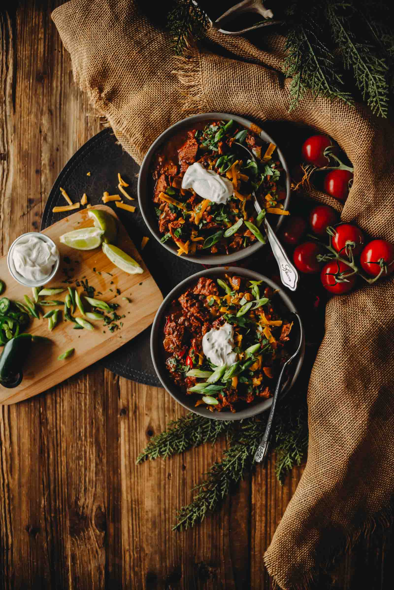 Two bowls of brisket chili topped with sour cream, cheese, and green onions on a burlap surface.