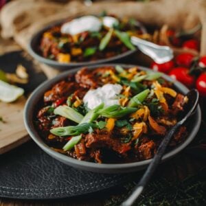 Bowl of brisket chili garnished with sour cream and sliced green onions, with a spoon resting on the side.