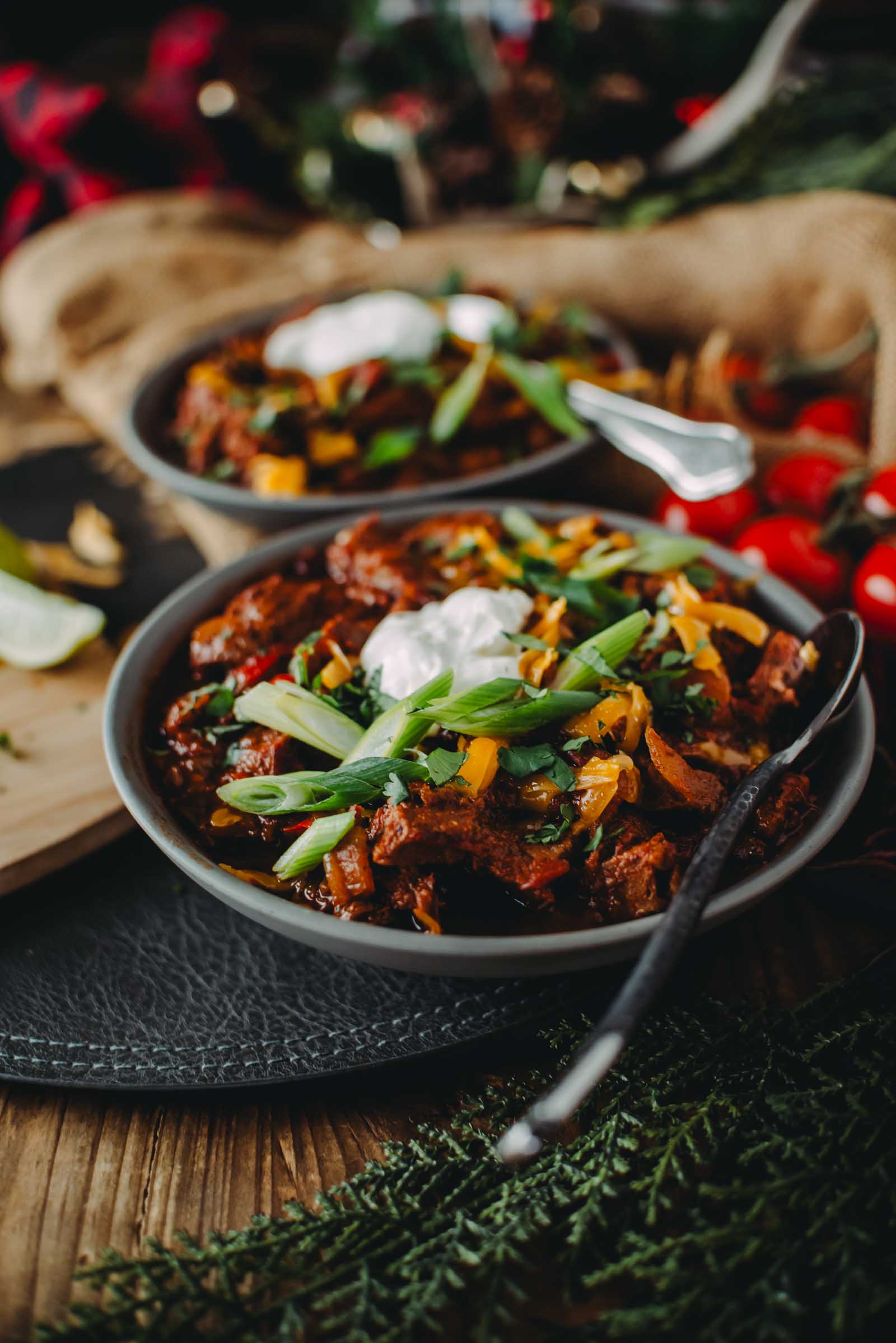 Bowl of brisket chili garnished with sour cream and sliced green onions, with a spoon resting on the side.