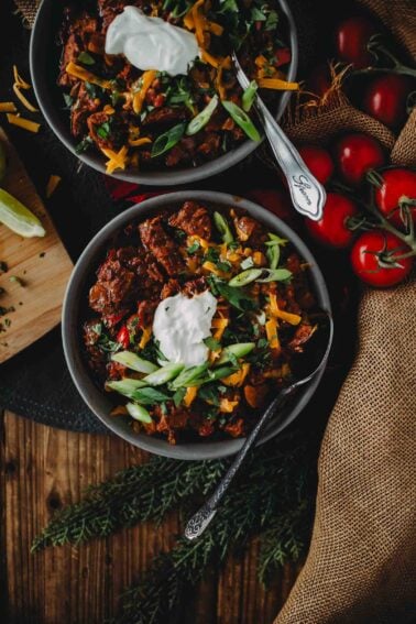 Two bowls of brisket chili topped with sour cream, cheese, and green onions, with a side of tomatoes, lime, and herbs on a rustic table setting.